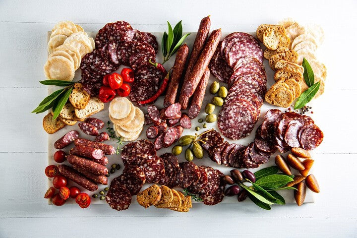 A deli platter showcasing a variety of salami slices, crackers, olives, and cherry tomatoes, elegantly arranged on a white plate.