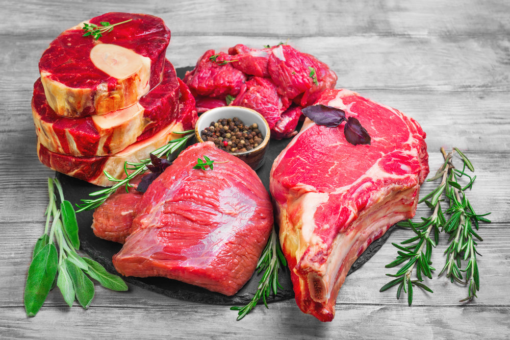 Different types of meat beef on round board. Herbs for an assortment of beef meat basil, thyme, rosemary, sage leaves, pepper. Light white wooden background. Top view from above.